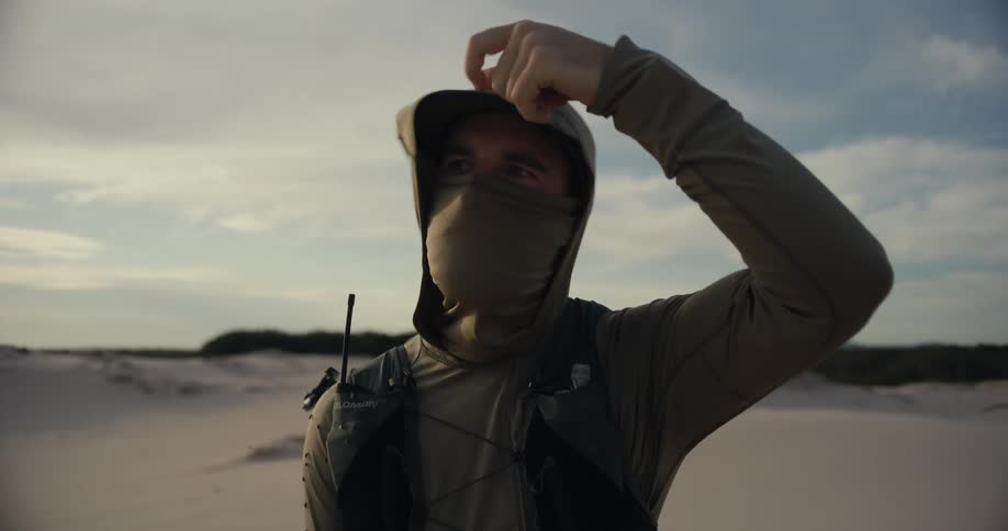 A solo adventurer in a desert landscape pulls up his neck gaiter and hood to protect himself from the elements in cinematic slow motion