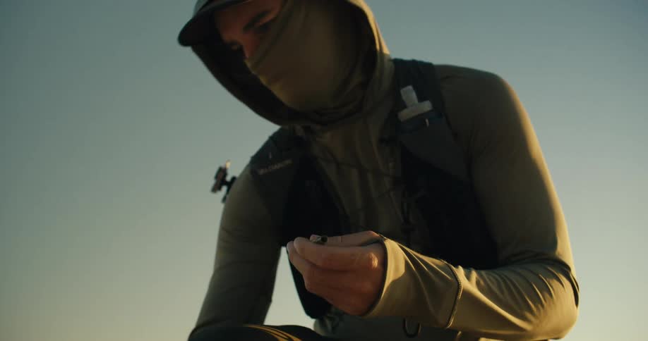 A hiker in a hoodie and face covering kneels down to inspect a bullet casing he found in the desert in slow motion