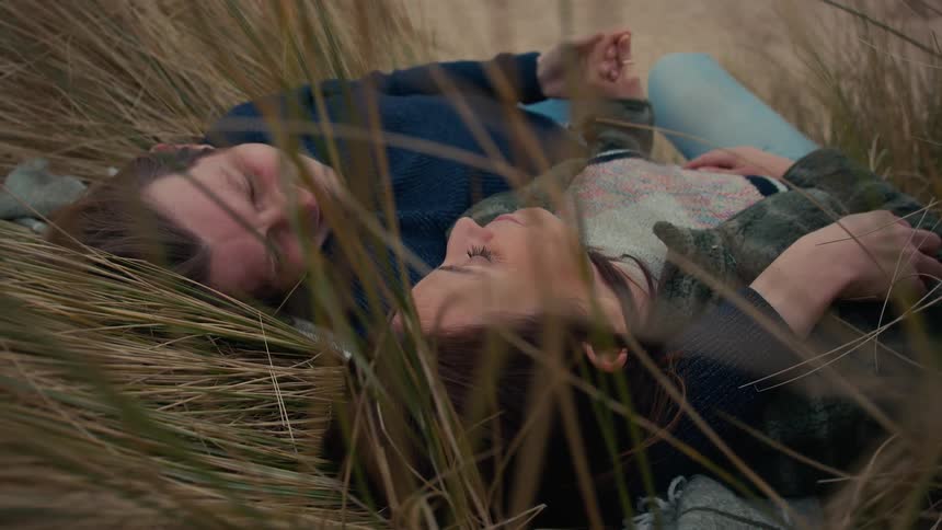 A loving couple lies peacefully together with their eyes closed, holding hands while nestled in the tall grass of a sand dune