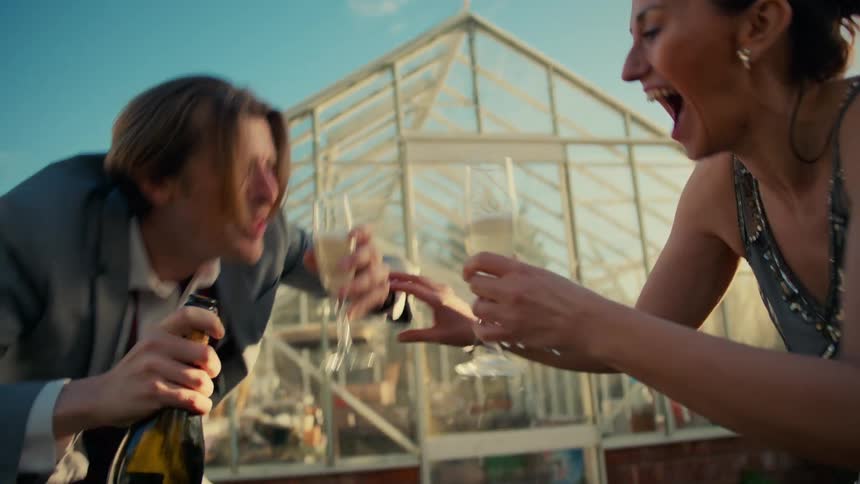 A playful couple has fun at an outdoor party, laughing hysterically as champagne overflows from their glasses in slow motion