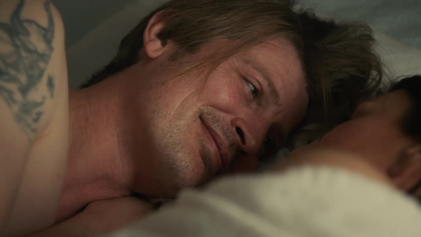 A loving man with a tattoo on his arm wakes up and smiles at his partner in bed
