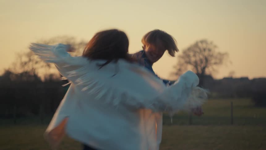 A happy young couple laughs and dances freely together in a grassy field during a beautiful golden hour sunset