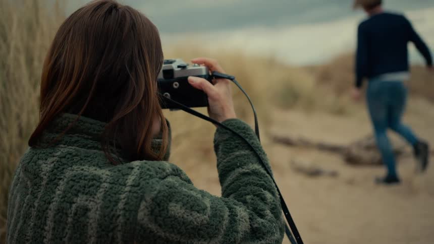 A smiling woman takes a picture of a man with a vintage film camera while sitting on a sandy dune