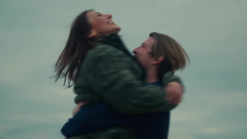 A joyful man lifts his girlfriend into the air and spins her around as they laugh and embrace in slow motion on a windy day