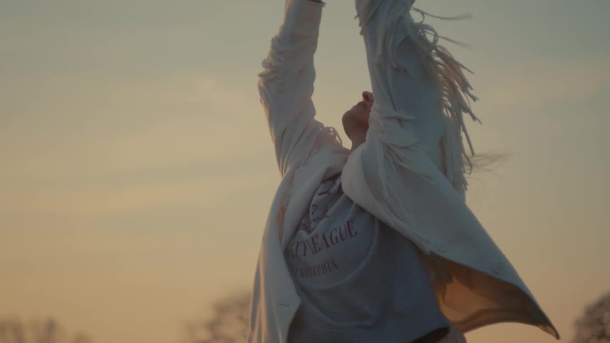 A young woman in a white fringed jacket stretches her arms to the sky in slow motion during a beautiful golden hour sunset