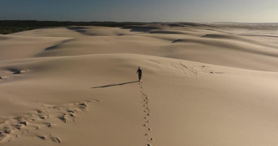 Aerial view of a lone traveler walking through a vast sand dune desert, leaving footprints in the golden light