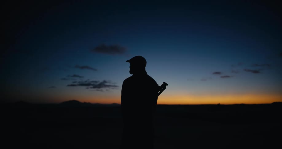 A lone hunter with a rifle is silhouetted against the dark blue and orange twilight sky