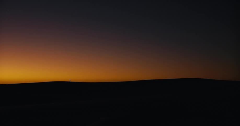 A lone silhouette of a person stands on a distant sand dune against the vibrant gradient of a sunset sky