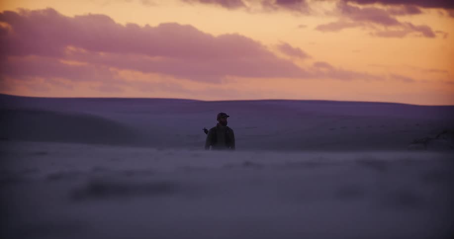 A lone adventurer walks through a vast, windy desert landscape under a beautiful purple and orange sunset