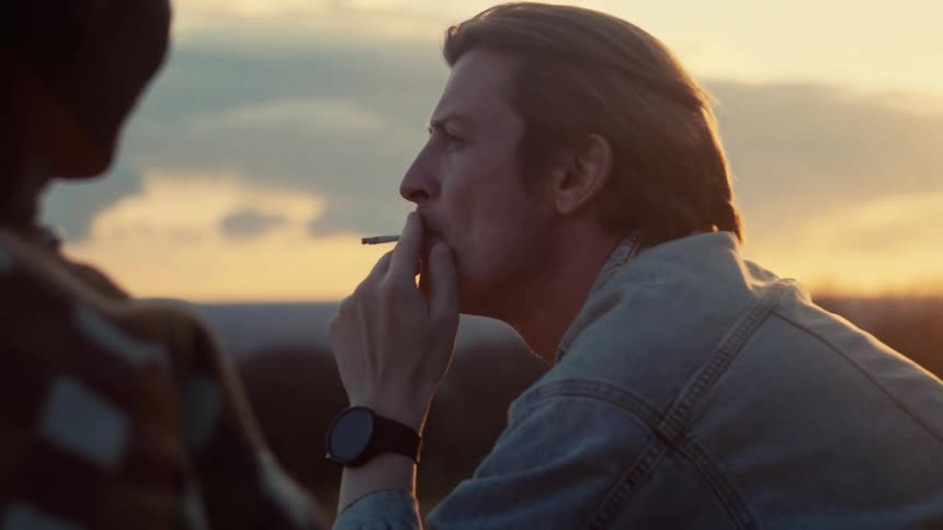 A man in a denim jacket shares a cigarette with a friend as they sit and talk during a beautiful sunset