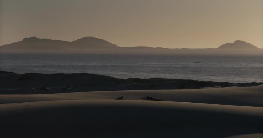 A serene, static shot of sand dunes overlooking a calm ocean with hazy islands on the horizon at sunset