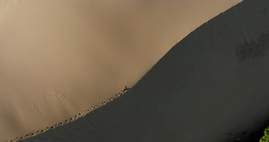 A high-angle top-down shot captures a lone person walking along the sharp ridge of a vast sand dune, leaving footprints behind
