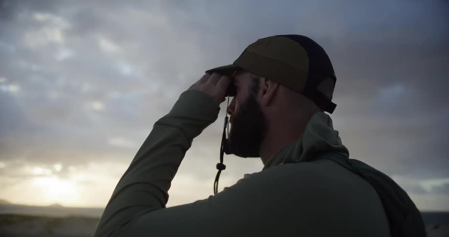 An outdoorsman with a beard and backpack uses a monocular to survey the coastal landscape at dusk