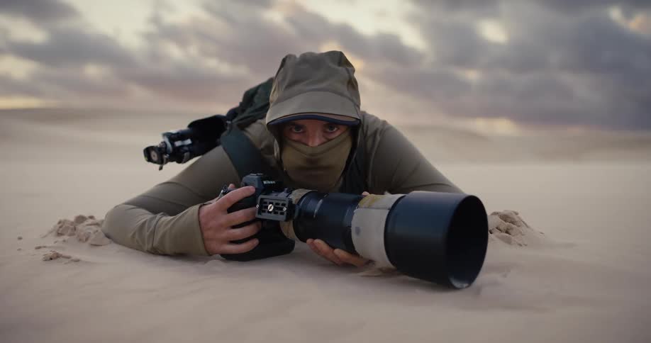 A dedicated nature photographer in full gear lies patiently on a desert sand dune with a large telephoto lens
