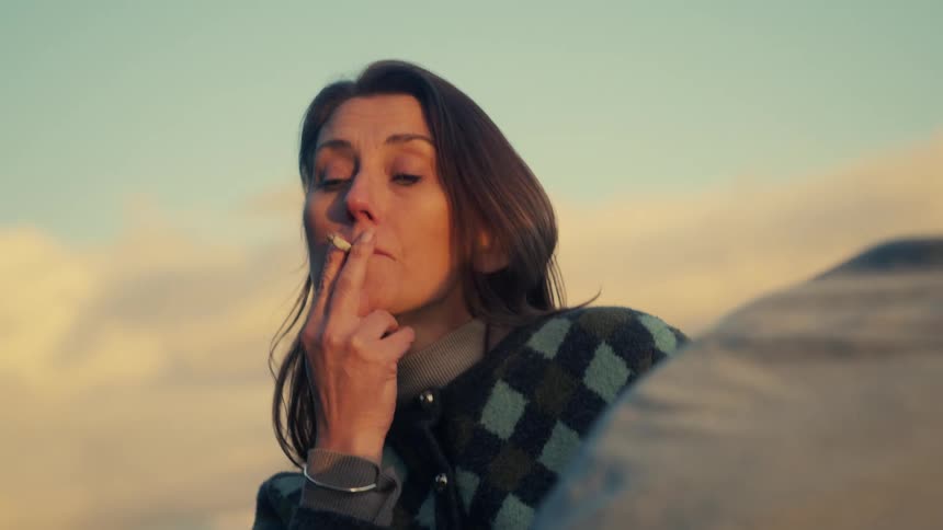 A woman in a cozy sweater has a violent coughing fit while smoking a cigarette and talking with a friend outdoors at sunset