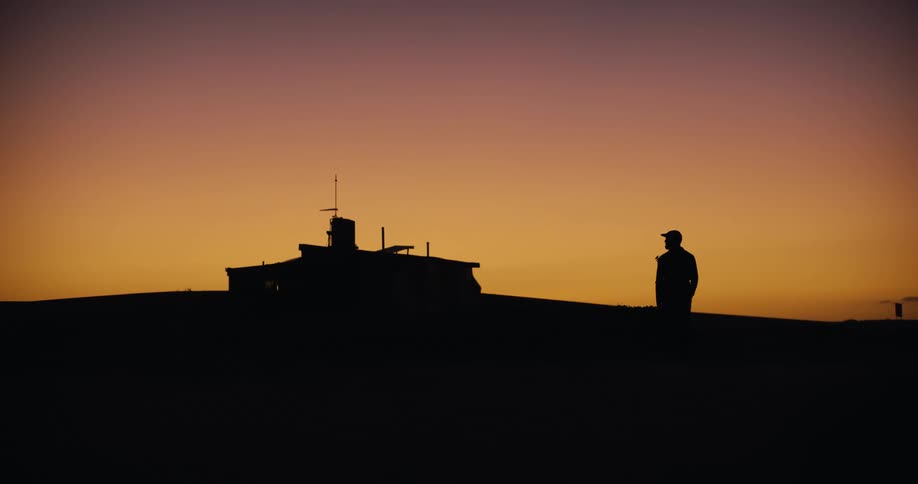 Silhouette of a lone man walking on a remote hilltop past a cabin against the vibrant colors of a sunset sky