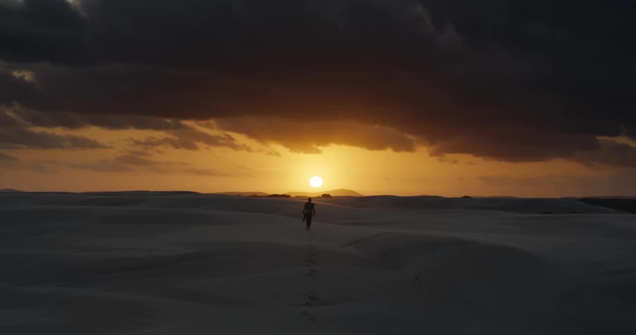 A silhouetted figure walks alone across endless sand dunes into the setting sun under a stormy sky