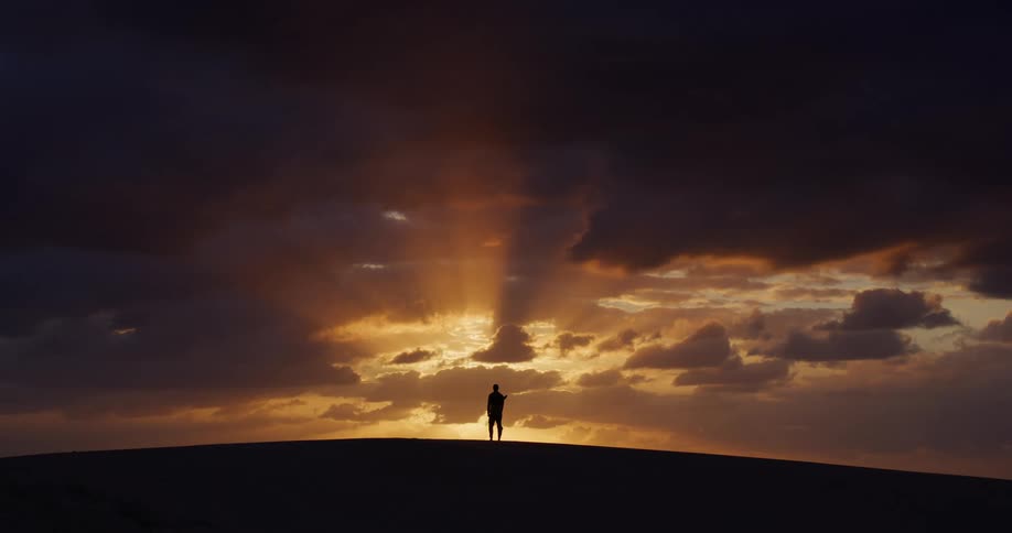 Silhouette of a lone figure standing atop a hill against a breathtaking sunset with golden light rays piercing through dark clouds