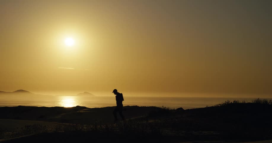 A silhouette of an adventurous photographer walks along a coastal ridge, overlooking the ocean during a beautiful golden sunset