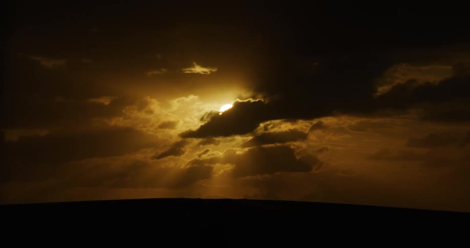 Sunbeams break through dark, heavy clouds during a dramatic golden hour sunset over a silhouetted landscape