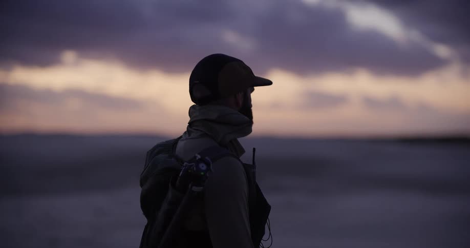 A solitary photographer with a tripod and backpack stands on the shore, observing the serene sunset over the ocean