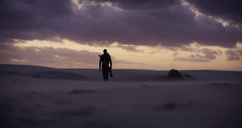 A lone photographer is silhouetted as he walks barefoot through a vast, sandy desert landscape towards the camera during a dramatic sunset