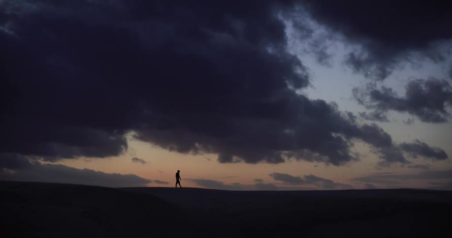 A lonely figure walks in silhouette across a vast desert landscape under a dramatic cloudy sky at dusk