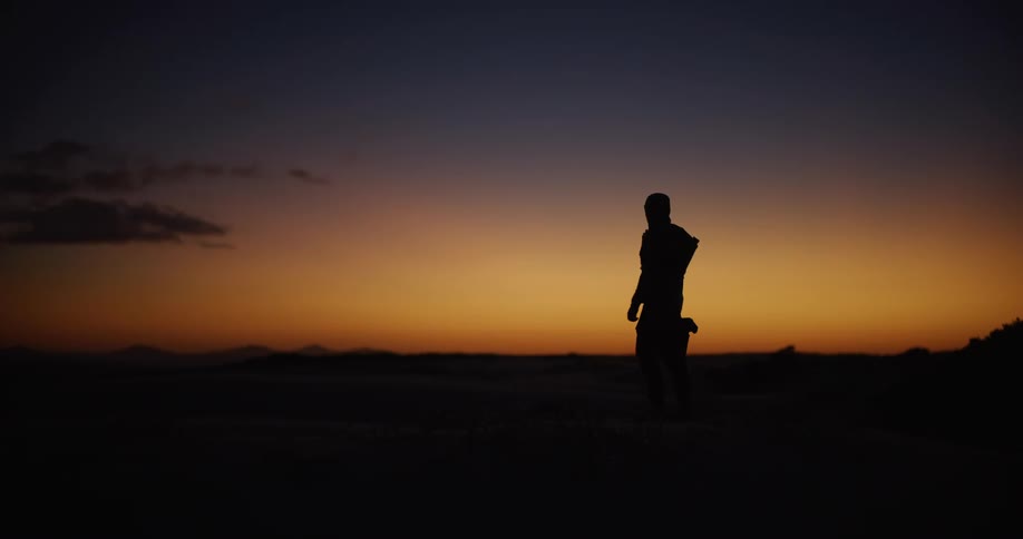 A silhouetted hiker walks into a desert landscape and pauses to stretch against the vibrant colors of a sunset sky