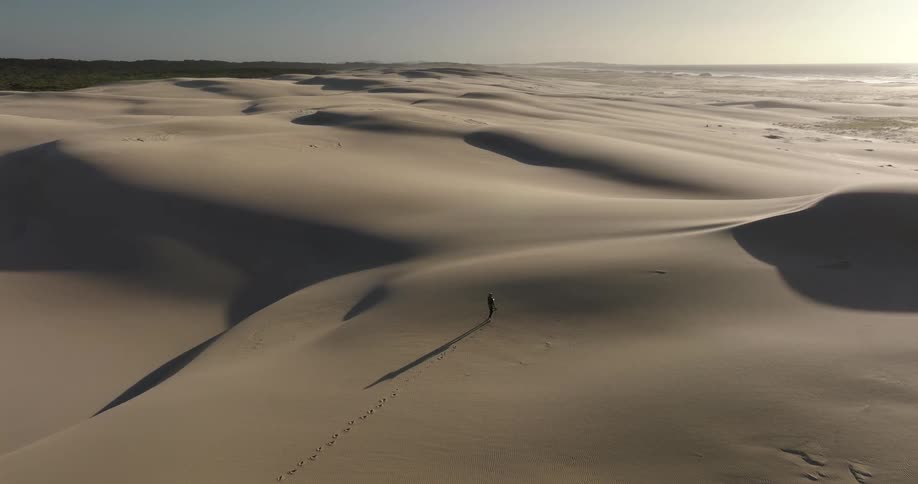 Aerial view of a lone person walking across vast, beautiful sand dunes at sunrise, leaving a trail of footprints behind