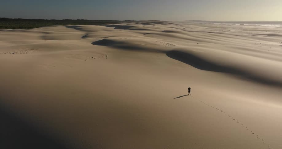 An aerial view shows a lone traveler walking across a vast expanse of sand dunes, leaving a trail of footprints behind them