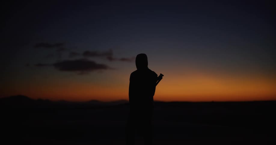 A silhouetted photographer with a tripod stands still in a vast, open landscape, enjoying the vibrant colors of the dusk sky