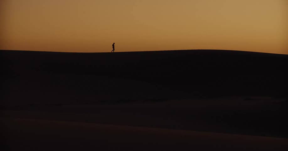 A silhouetted figure walks along the crest of a large sand dune against a beautiful orange sunset
