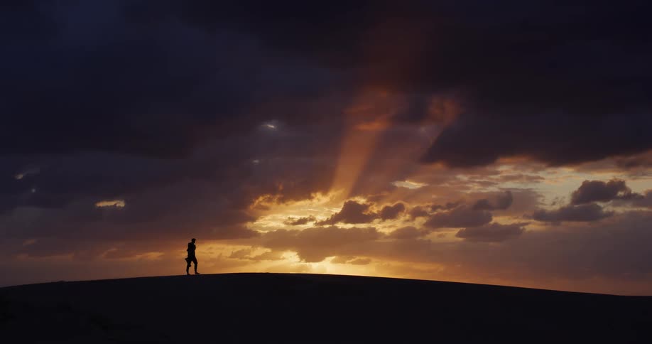 A lone traveler walks in silhouette along a vast sand dune against a dramatic, beautiful sunset with golden rays