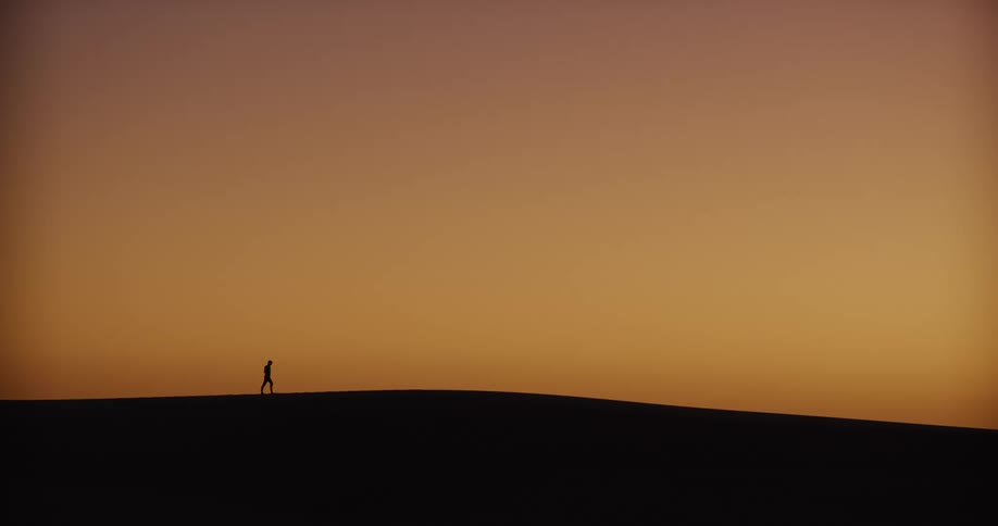 A solitary figure walks along the horizon of a vast desert at dusk and pauses to take in the view