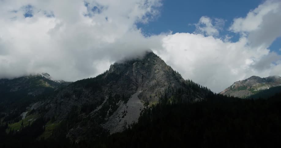Clouds drift over a rugged mountain peak in a dramatic, sun-dappled landscape