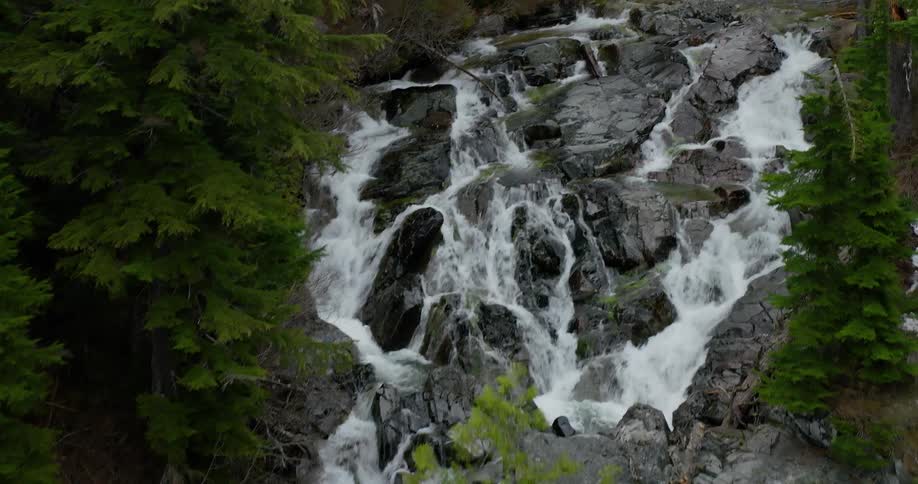 An aerial view ascends a beautiful cascading waterfall flowing over rocks and through a dense evergreen forest in the mountains