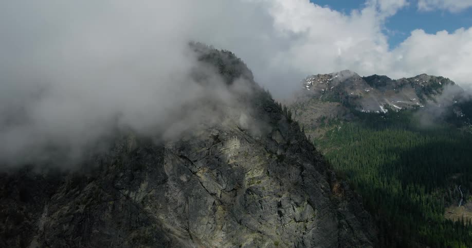 A dramatic aerial shot tilts up from a dark, rocky mountain face, revealing a vast, cloud-covered mountain range with lush green forests
