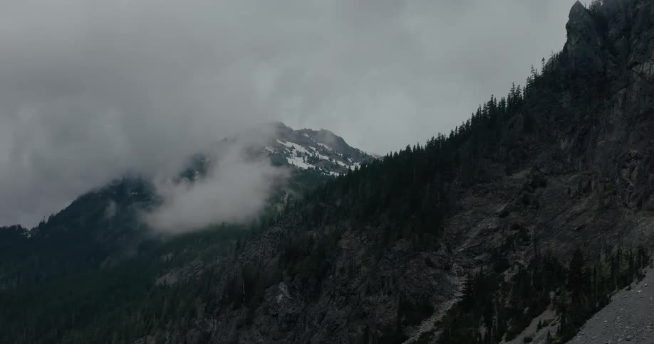Aerial view slowly pulls back from a dramatic, moody mountain range with snow-capped peaks shrouded in thick fog and clouds