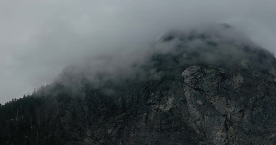 Slow aerial dolly shot moving towards a rugged mountain peak shrouded in thick, moody fog