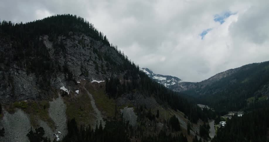 An aerial view pulls back over a vast, rugged mountain range covered in pine forests and patches of snow under a dramatic, cloudy sky