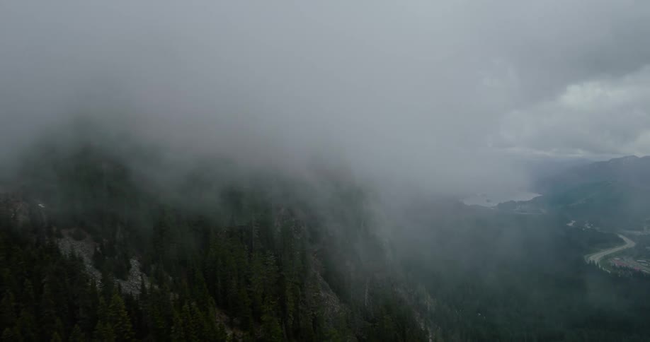 An aerial view pulls back from a dramatic, fog-covered mountain peak to reveal a vast evergreen forest and a valley with a lake below