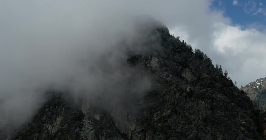 A dramatic aerial shot slowly pulls back from a majestic, cloud-covered mountain peak, revealing the surrounding evergreen forest