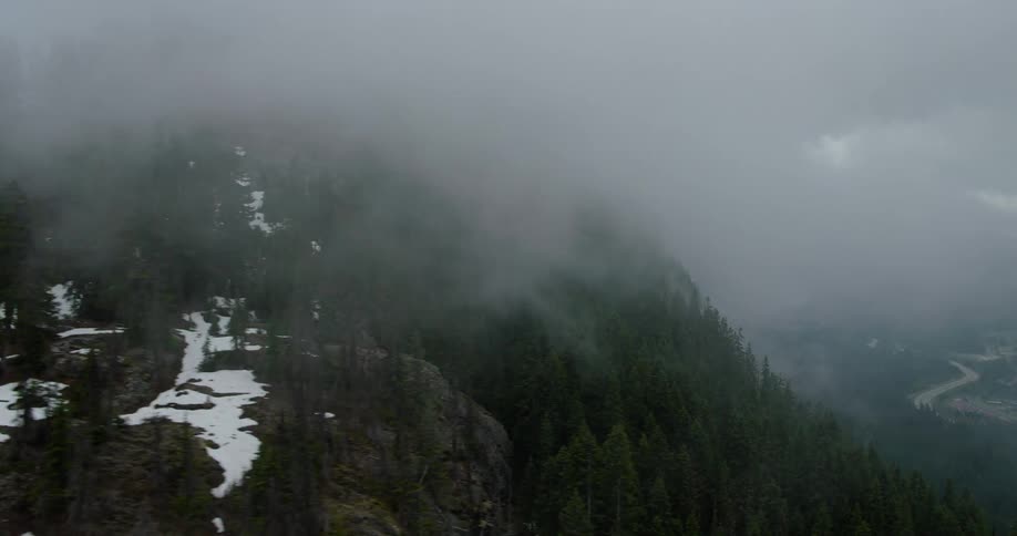 A dramatic aerial view pans across a rugged mountain peak shrouded in thick fog and clouds, with a lush pine forest below