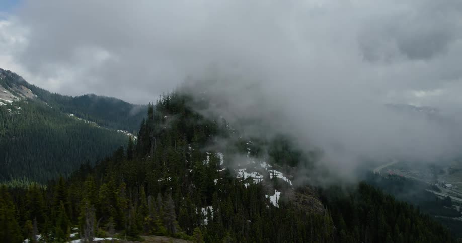 An aerial shot slowly ascends revealing a majestic, cloud-shrouded mountain peak covered in a lush evergreen forest