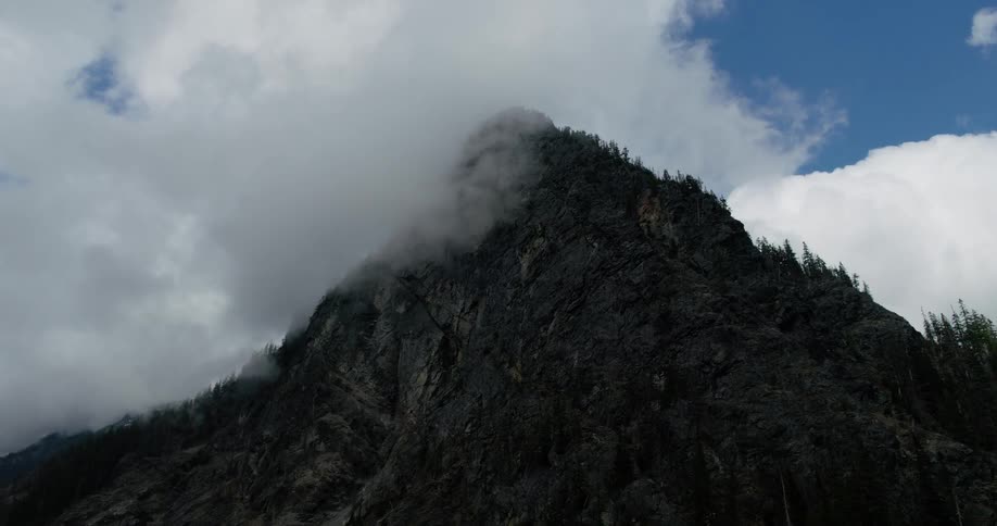 Moody, slow-moving clouds drift across a rugged, dark mountain peak against a blue sky
