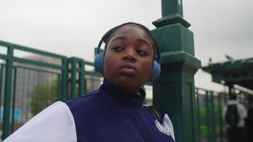 A thoughtful young Black woman wearing headphones listens to music while waiting at a train station platform