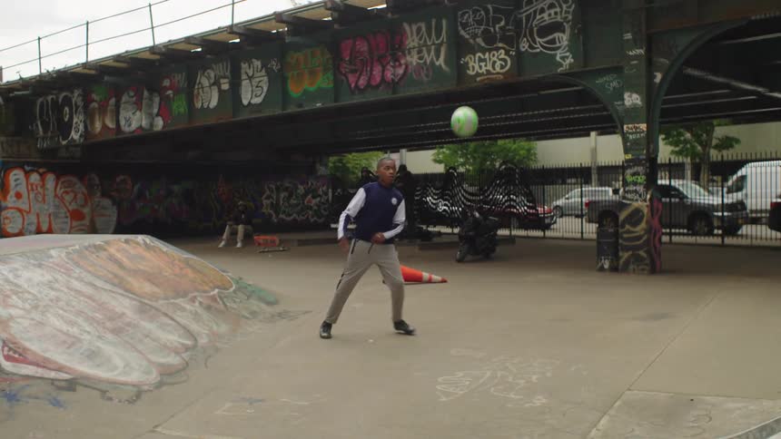 A young boy and a man in tracksuits practice their soccer juggling skills in slow motion at a graffiti-covered urban skatepark
