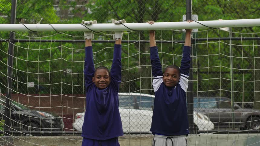 Two smiling young boys hang playfully from the crossbar of a soccer goal on a turf field in a city park