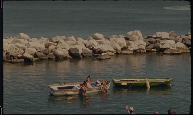 Friends relax and sunbathe on a small wooden boat in a calm Mediterranean bay in this vintage film footage
