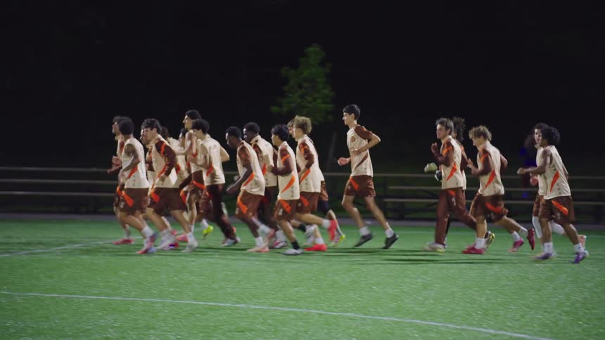A diverse men's soccer team runs together in formation across a brightly lit turf field during a night training session